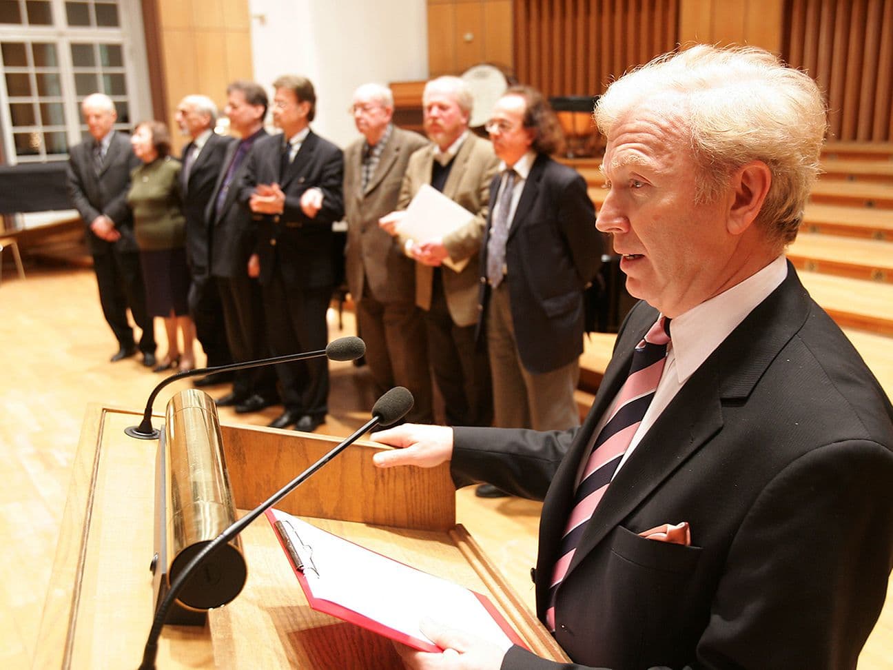 A man in a suit is giving a speech behind a lectern, while a group of seven people stand in the background.