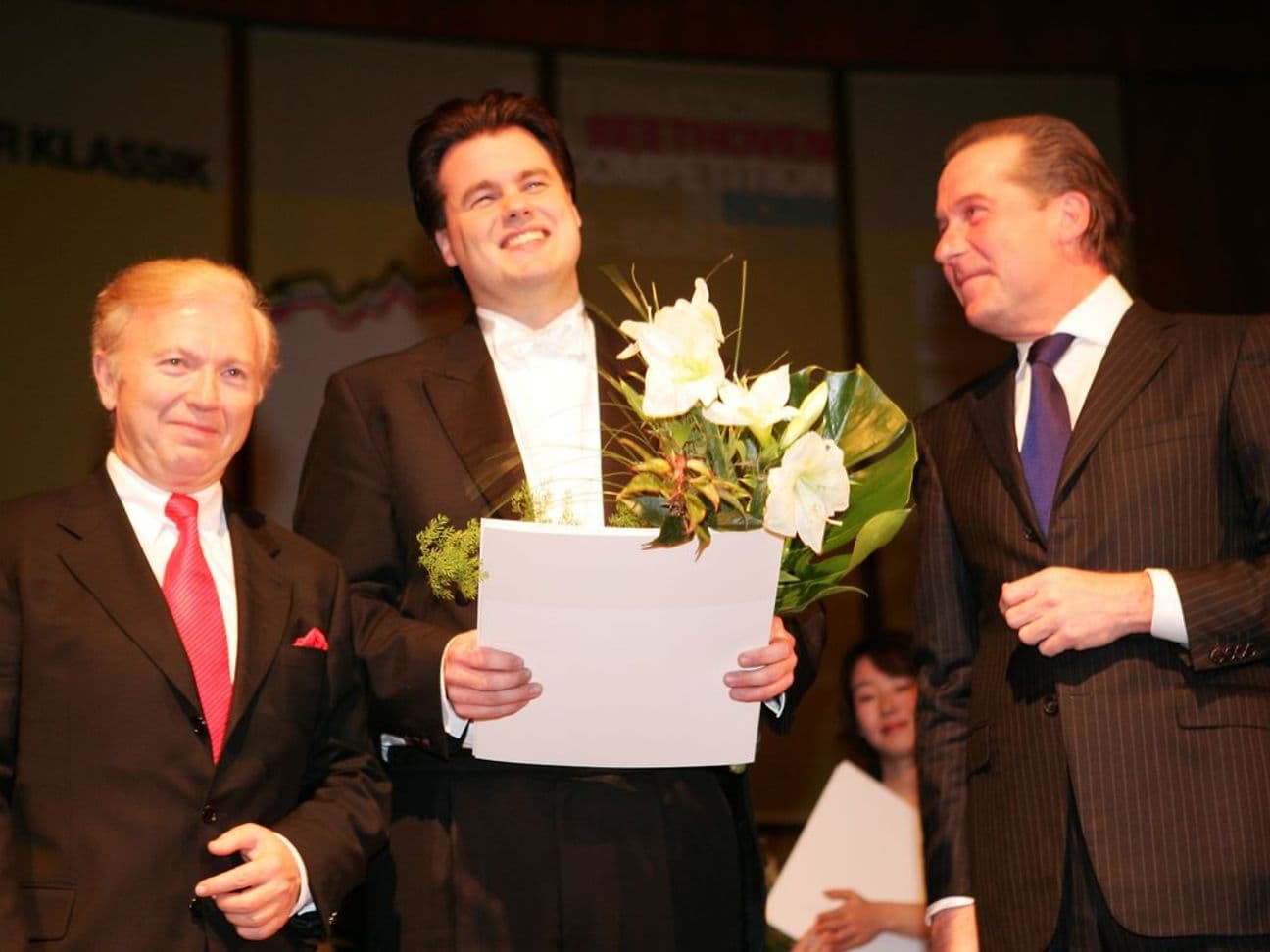 Three men in suits standing on a stage. The man in the middle is holding flowers and a paper, smiling.