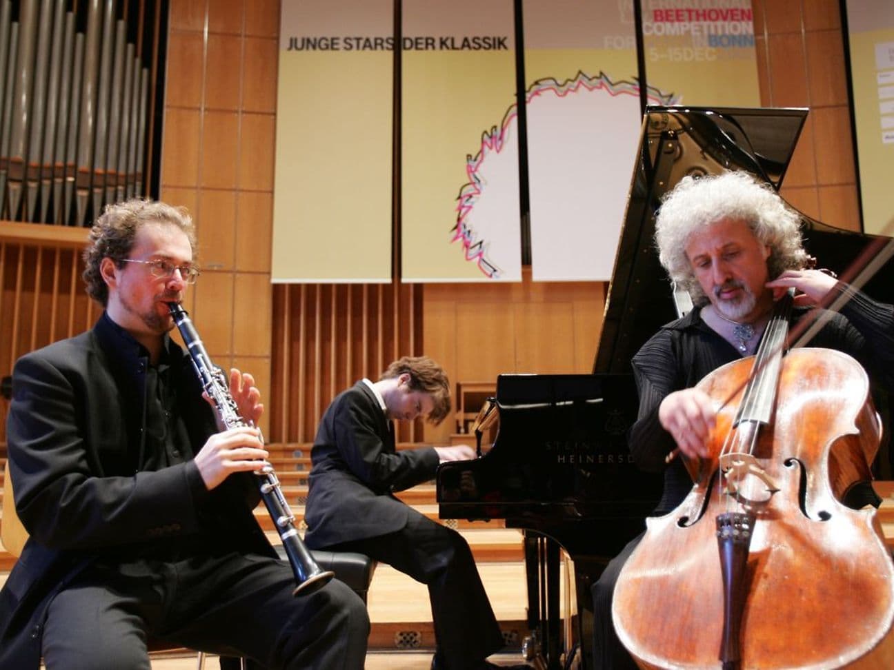 Drei Musiker spielen klassische Musik, ein Klarinettist, ein Pianist und ein Cellist. Im Hintergrund steht ein Schild mit der Aufschrift 'Junge Stars der Klassik' und das Wort 'Beethoven'.