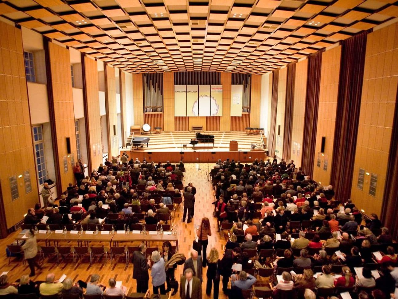 A fully occupied concert hall with a piano on the stage.