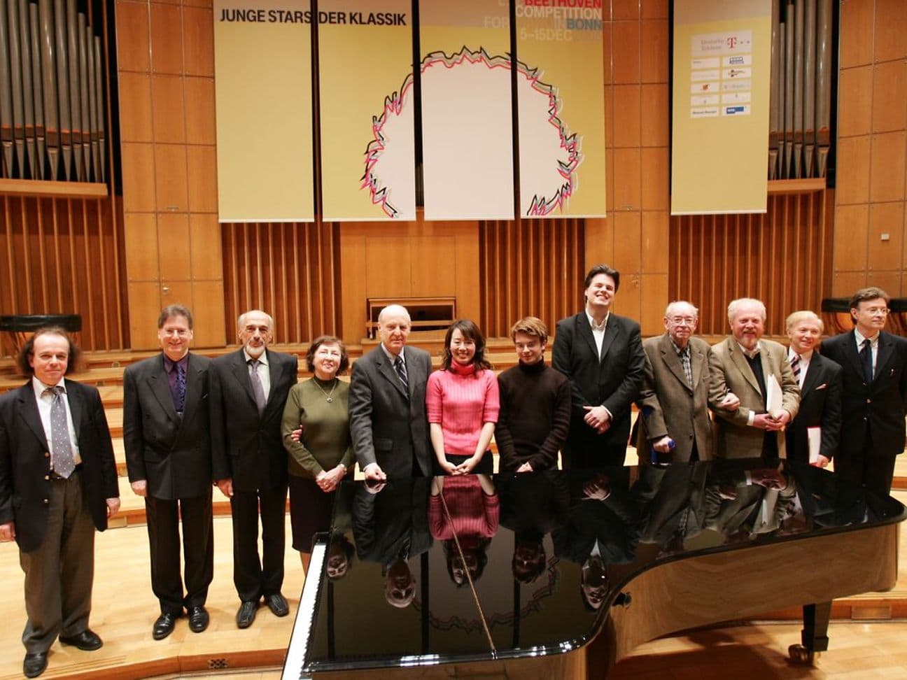 A group of people stand in front of a grand piano on a stage. In the background, a banner reads 'Young Stars of Classical Music'.