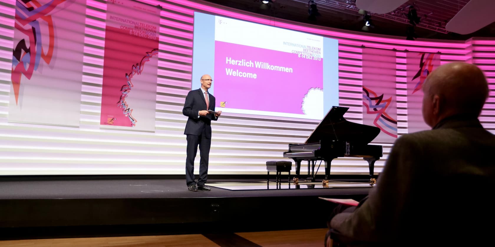 A man is giving a presentation on the stage of a concert hall. The screen reads 'Herzlich Willkommen - Welcome' at the International Telekom Beethoven Competition Bonn 6-14 December 2013. A grand piano is visible in the background.