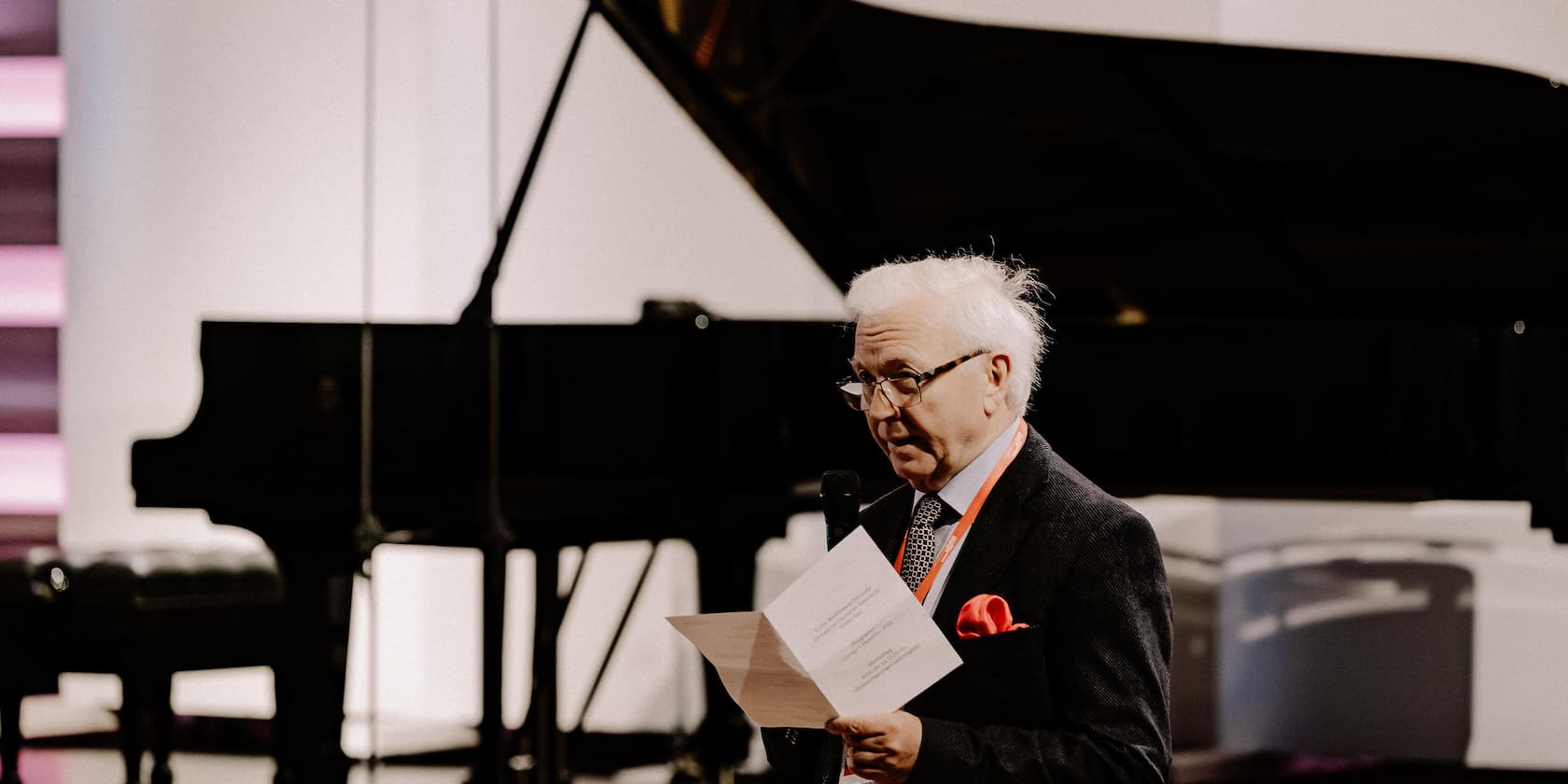 Elderly man with a microphone reads from a sheet of paper in front of a piano.