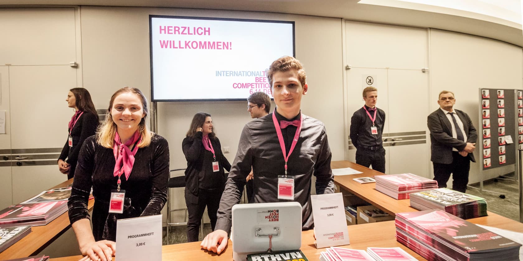 Several people are standing at tables with flyers and brochures at an event. In the background, there's a welcome screen with the text 'Herzlich Willkommen!'.