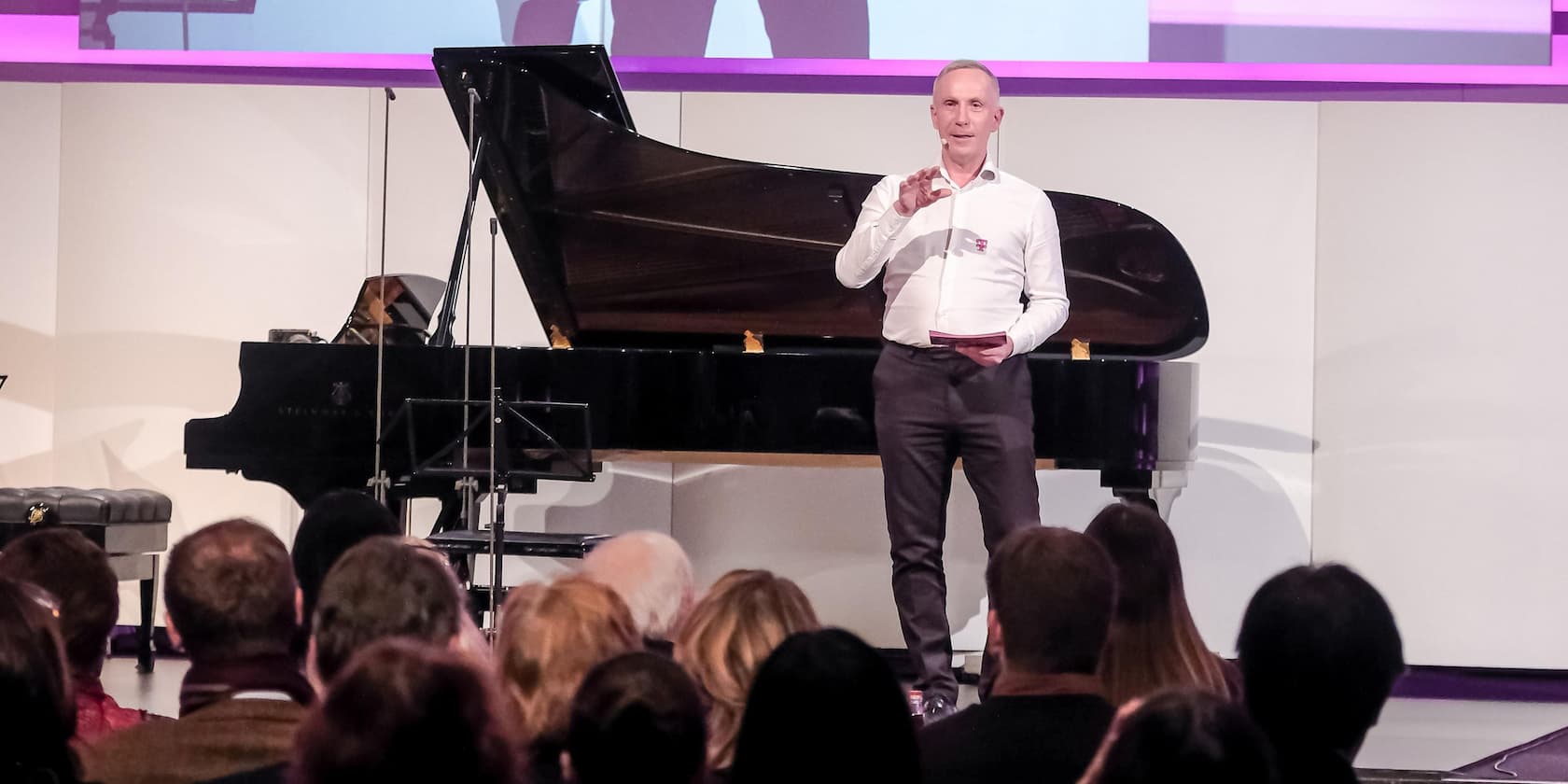 A man is giving a presentation to an audience with a grand piano in the background.