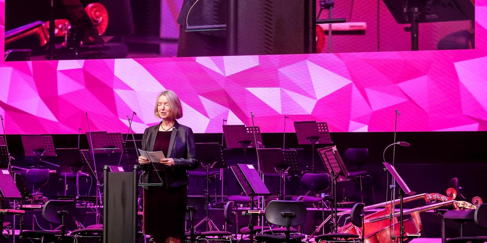 A woman is giving a speech in front of an orchestra with a large pink screen in the background.