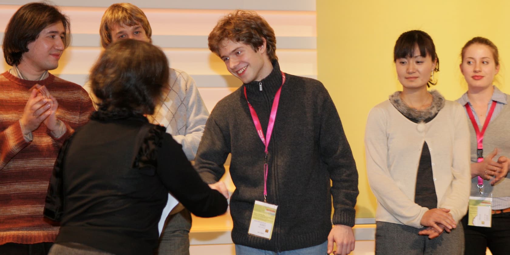 A group of six people standing on a stage. A man shakes a woman's hand while two people on the left applaud and two people on the right look on.