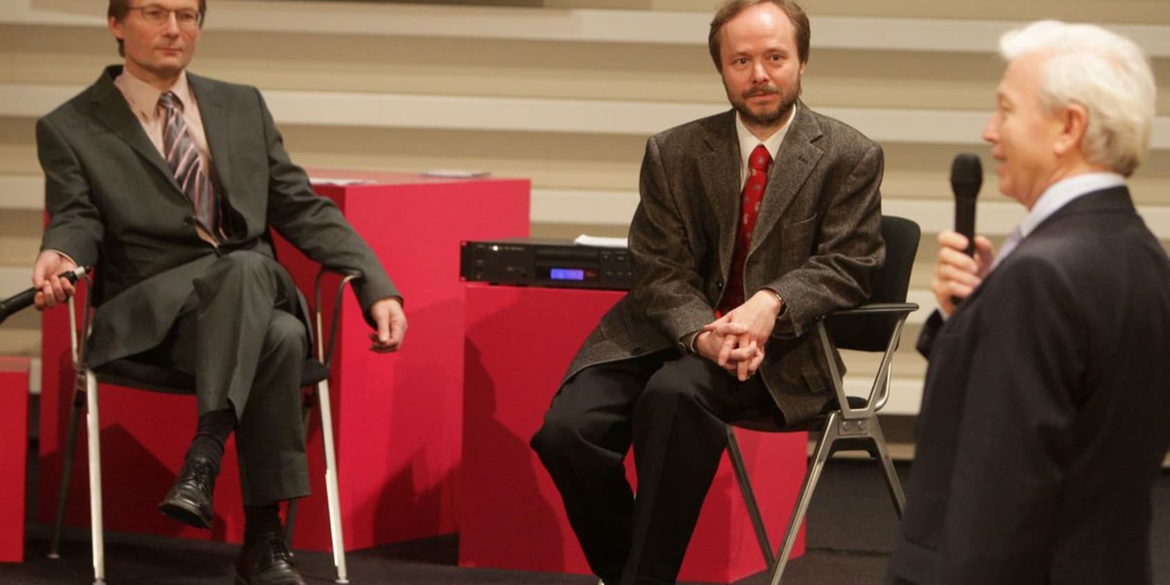 Three men in suits having a discussion in a room; one is standing with a microphone, the other two are seated.