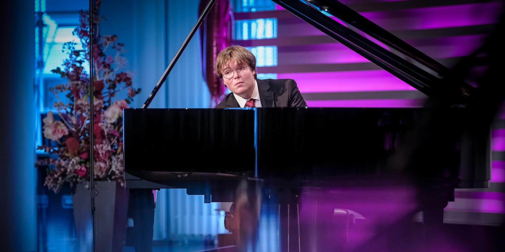 A man in a suit playing the piano in a room with colorful lighting and a flower arrangement.