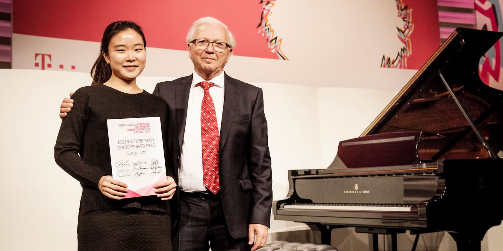 A woman holds a certificate reading 'Best Interpretation Contemporary Piece' beside an older man in front of a grand piano.