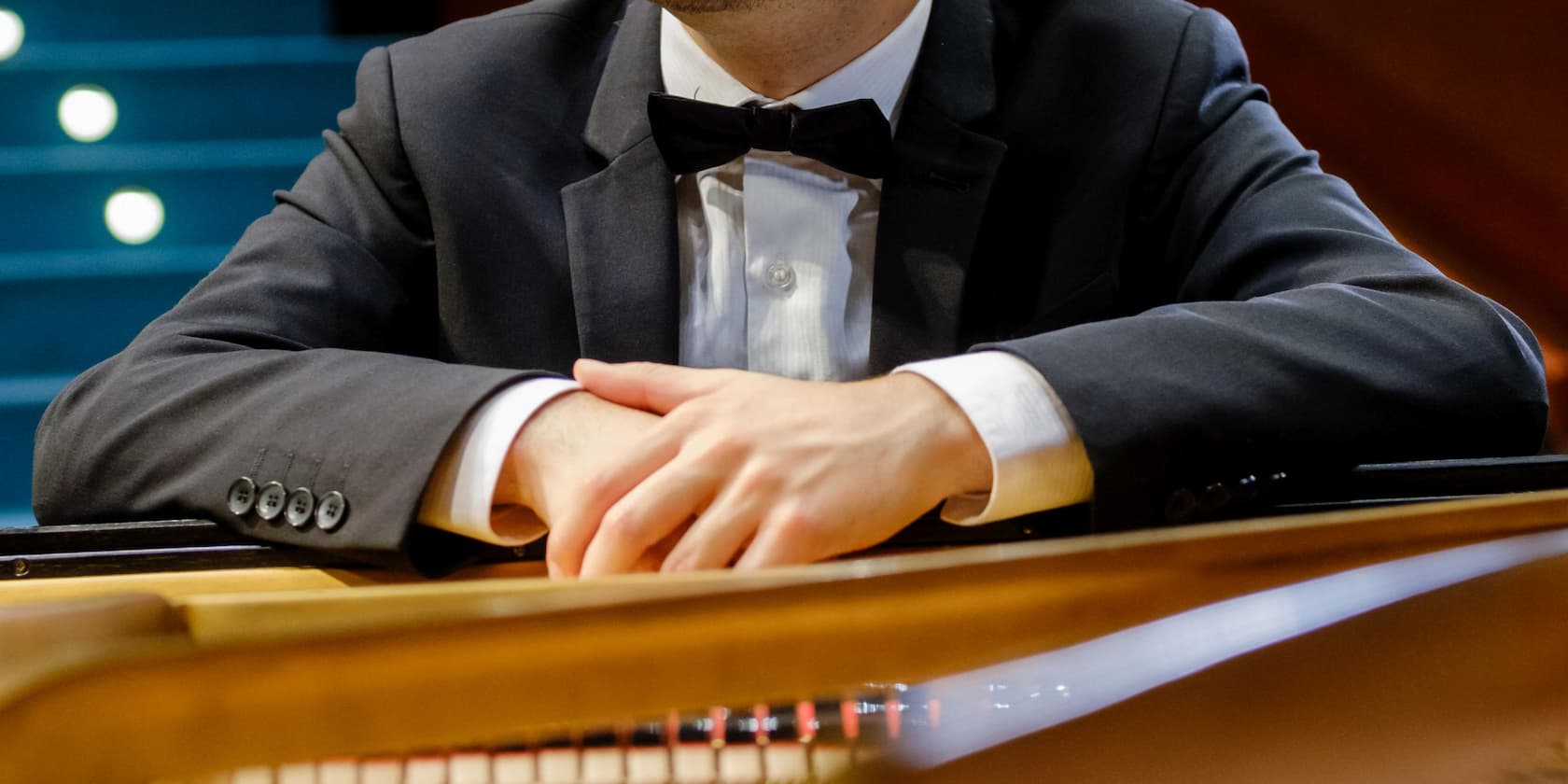 Close-up of a man in a suit with a bow tie, resting his arms on a piano.