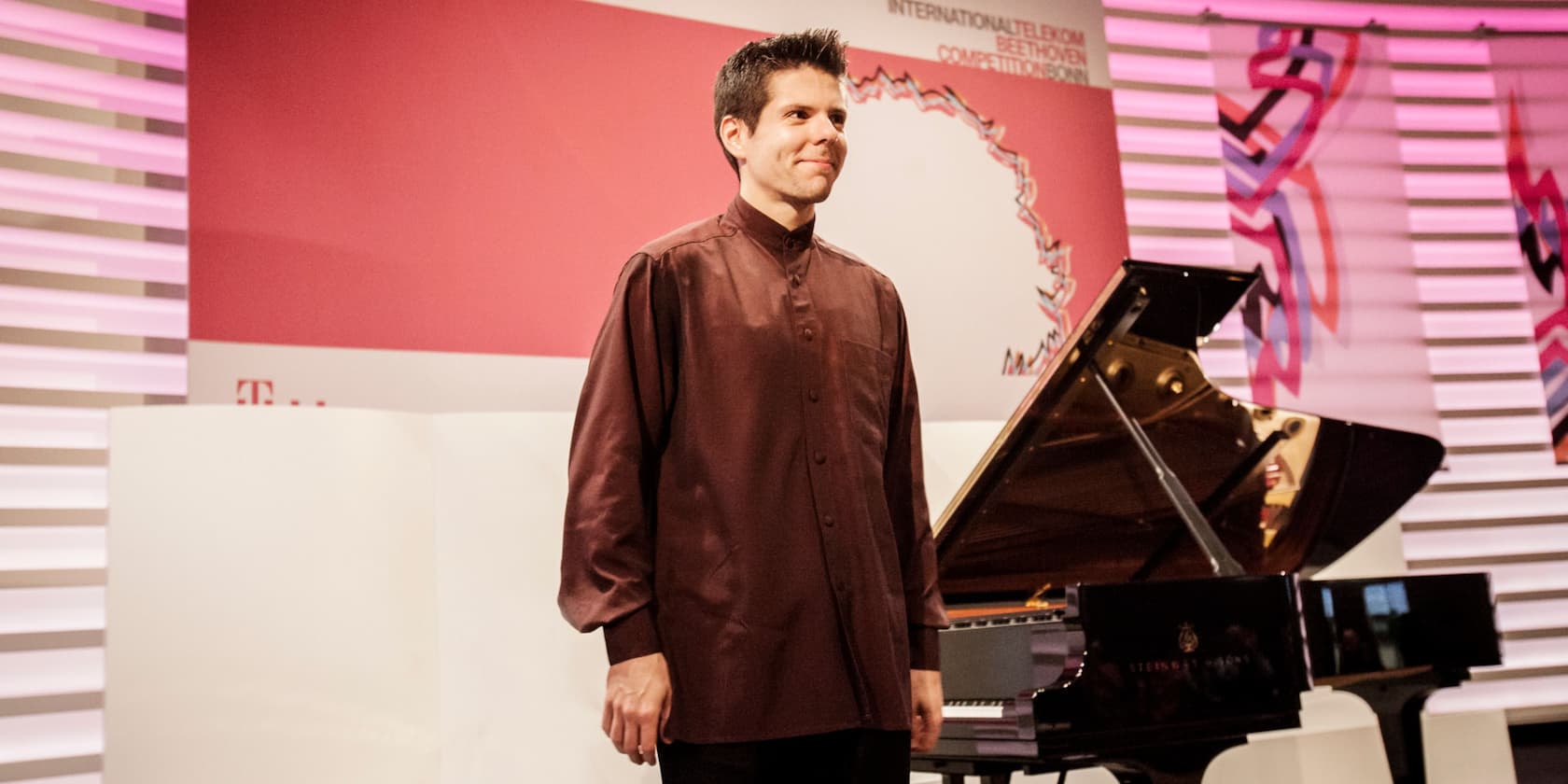 A man in a brown shirt stands in front of a grand piano during the International Telekom Beethoven Competition.