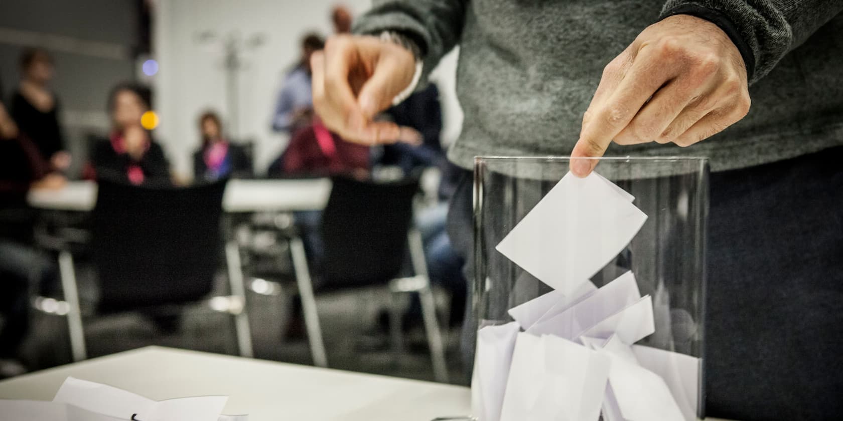 Hand legt einen Zettel in eine Wahlurne, Personen im Hintergrund.