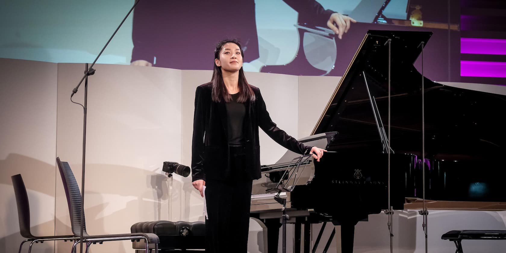 A woman stands next to a grand piano on a stage.