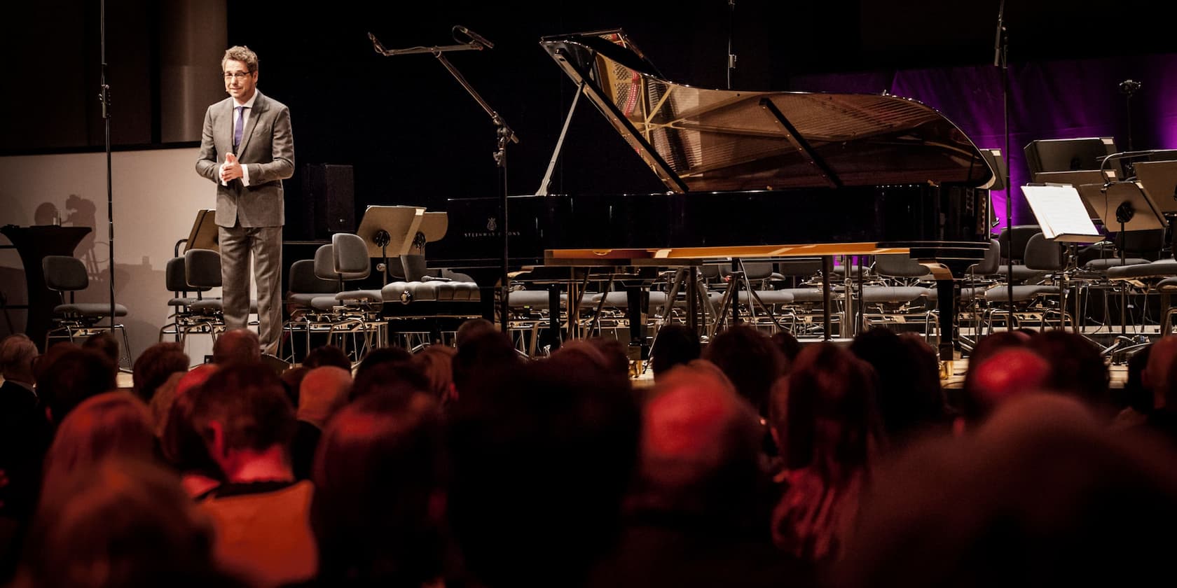 A man standing on a stage speaking to an audience. In the background, there is a grand piano and empty chairs.