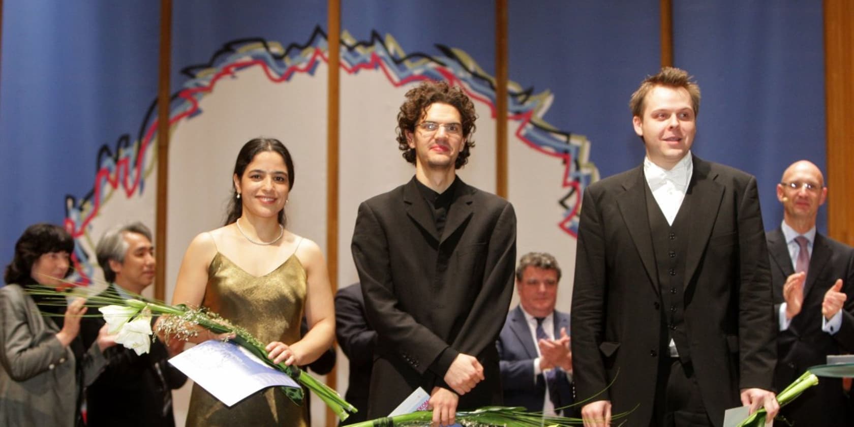 Three people standing side by side on a stage, holding flowers and certificates, while people in the background applaud.