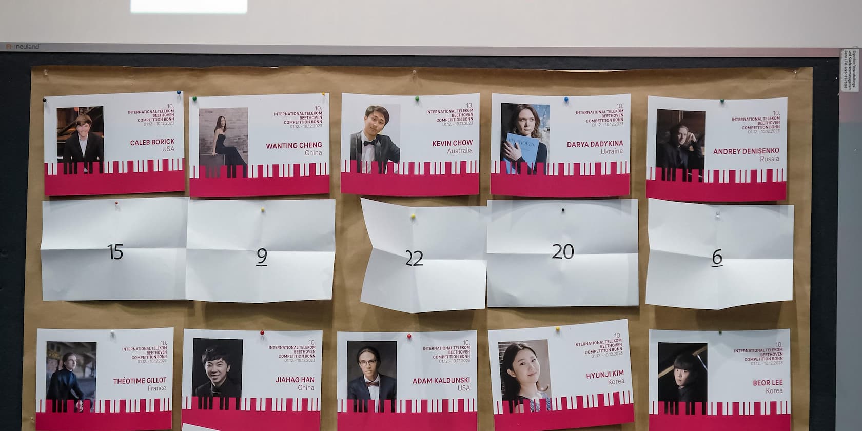 Wandtafel mit Teilnehmenden des 10. Internationalen Telekom Beethoven Wettbewerbs in Bonn. Bilder und Namen der Teilnehmer sind auf Papier gedruckt.