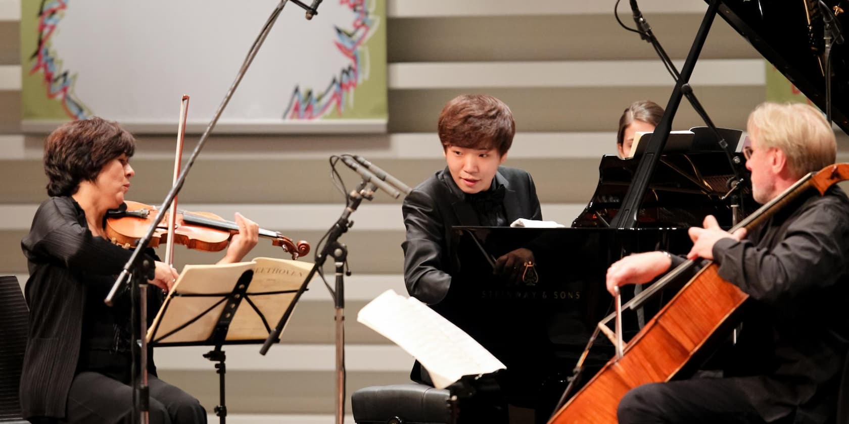 Three musicians playing classical music: a woman with a violin, a man at the piano, and a man with a cello.