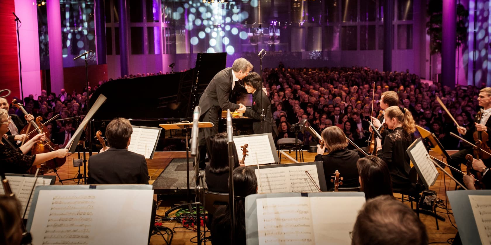 The conductor greets the pianist on stage during a concert in a fully packed hall, with the orchestra arranged around them.