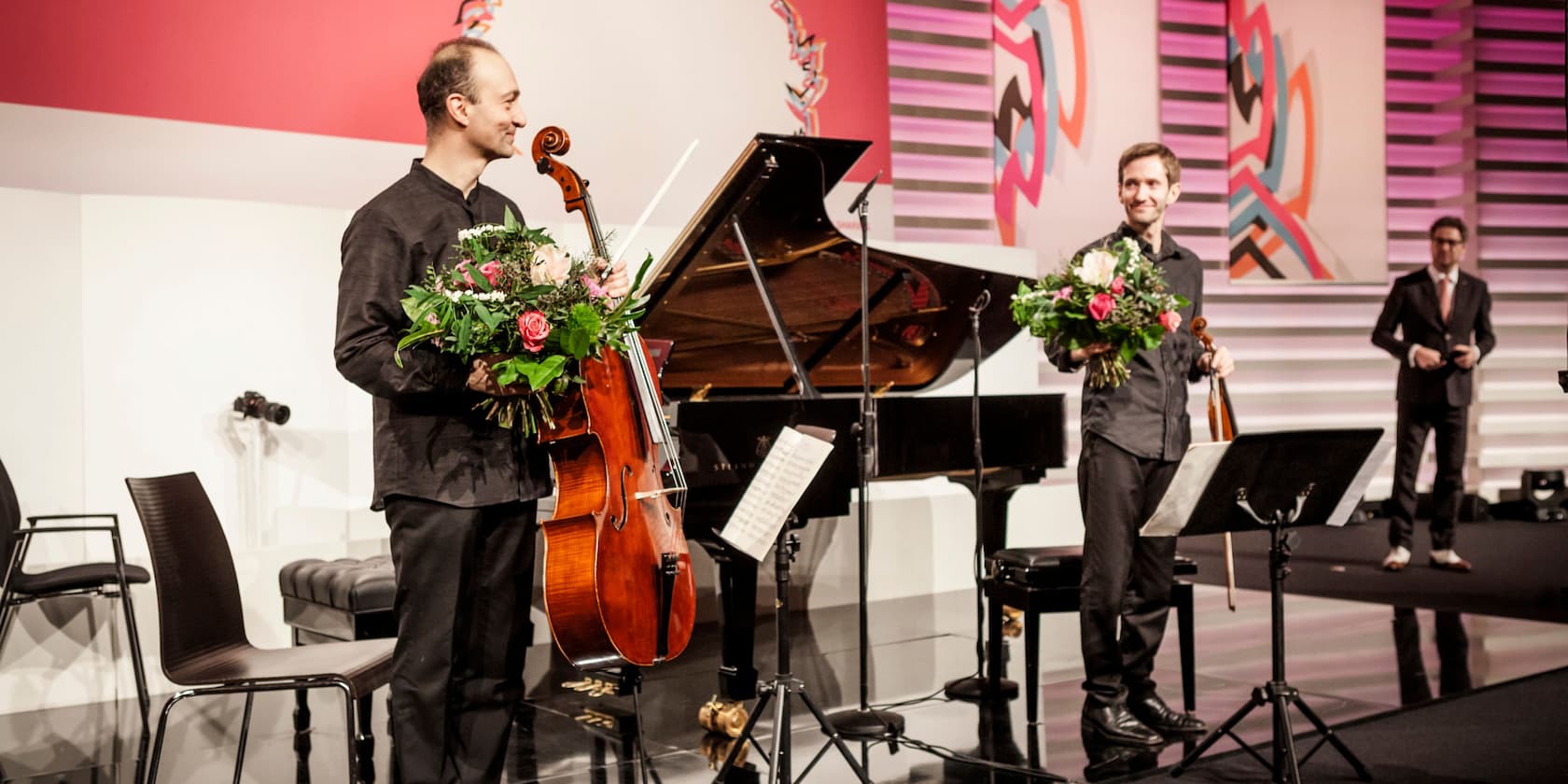 Two musicians with bouquets of flowers stand next to a piano and a cello on a stage.