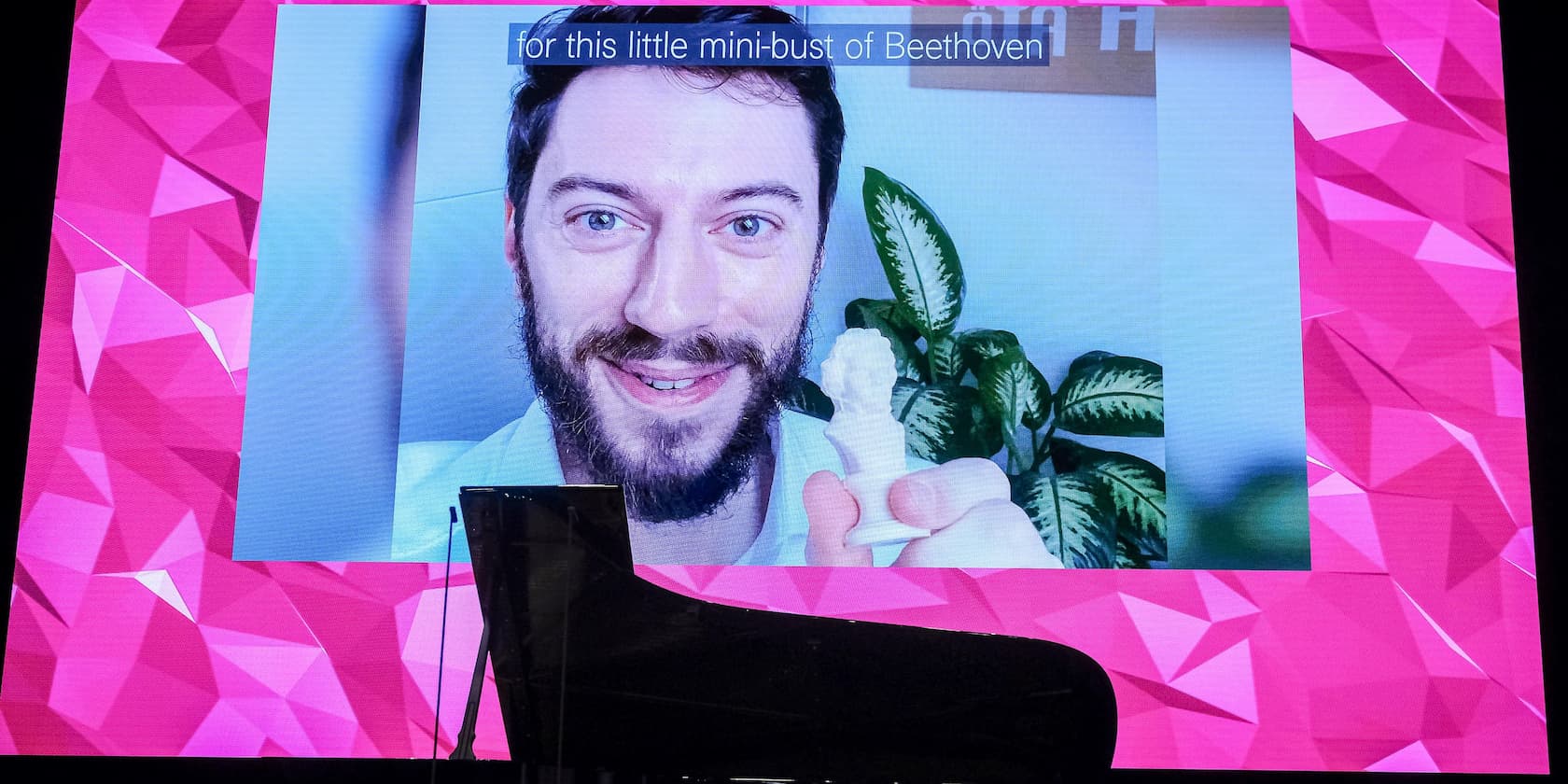 A bearded man holding a small bust of Beethoven is displayed on a screen. In the foreground, a black grand piano is visible.