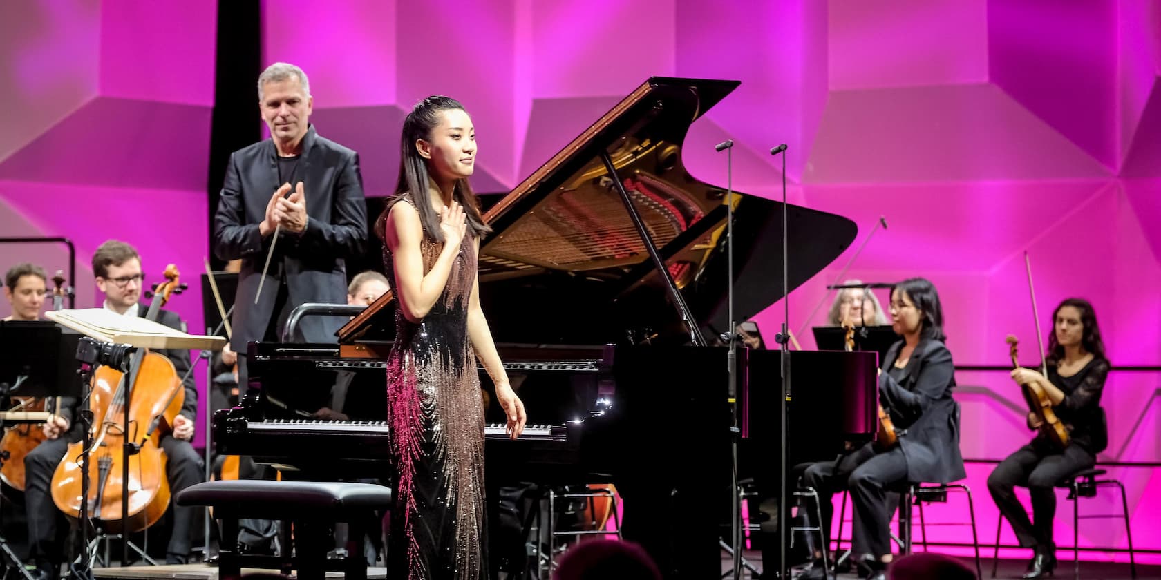 A pianist stands next to a grand piano after her performance while a conductor and an orchestra applaud. The background is illuminated with vibrant pink lighting.