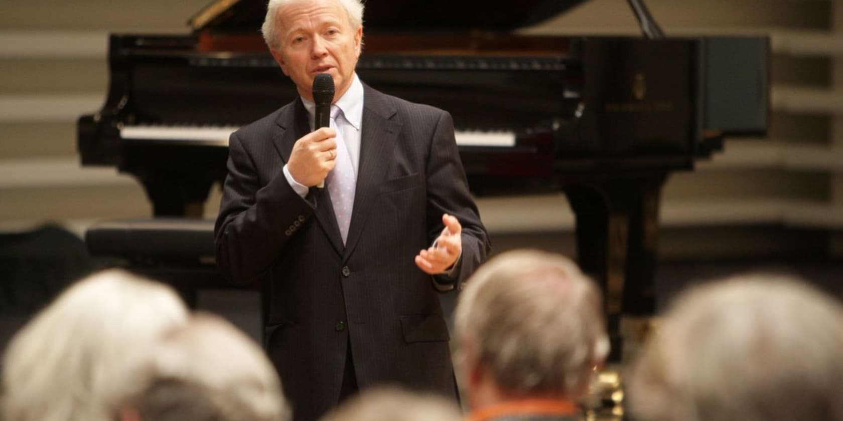 An elderly man giving a speech with a microphone in front of a grand piano.