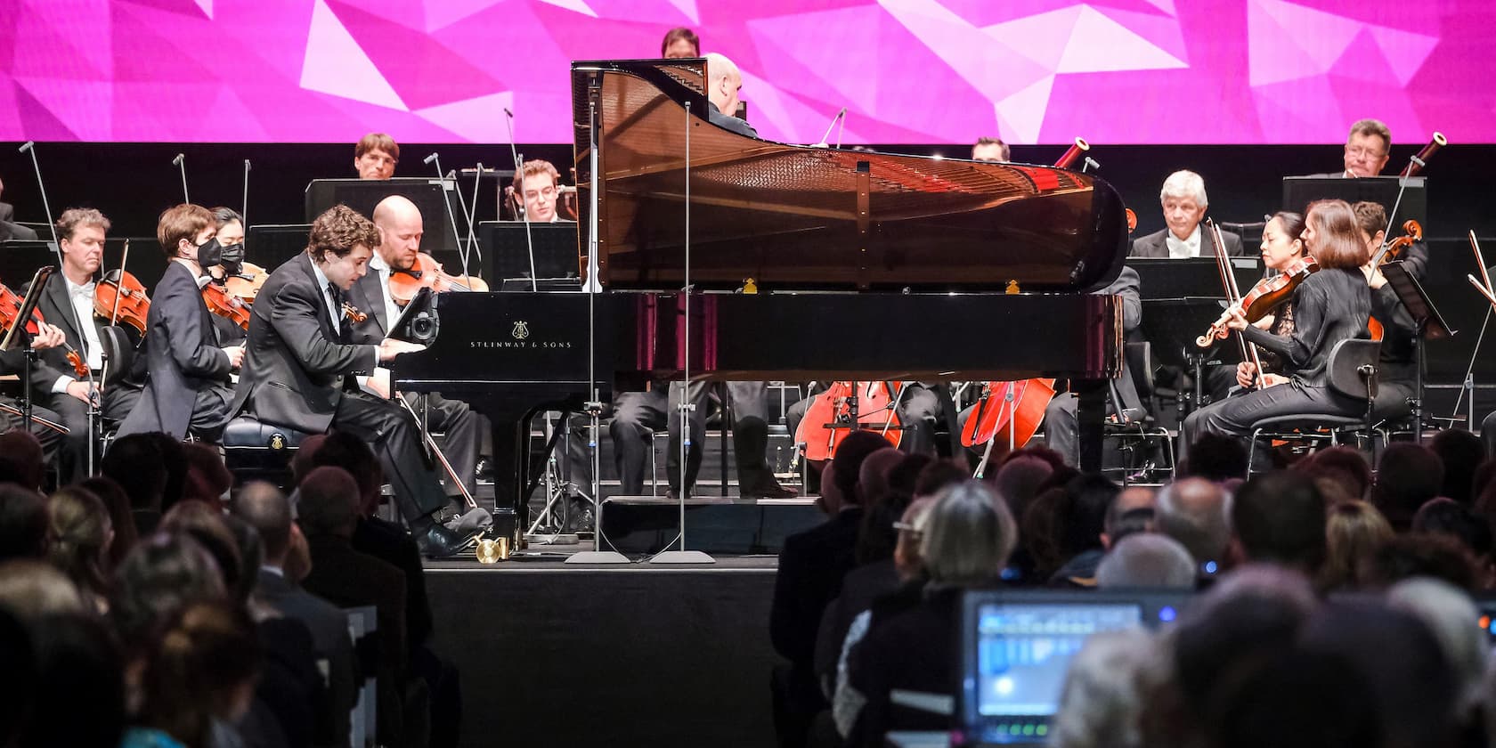 Ein Pianist spielt mit einem Orchester auf einer Bühne vor Publikum.