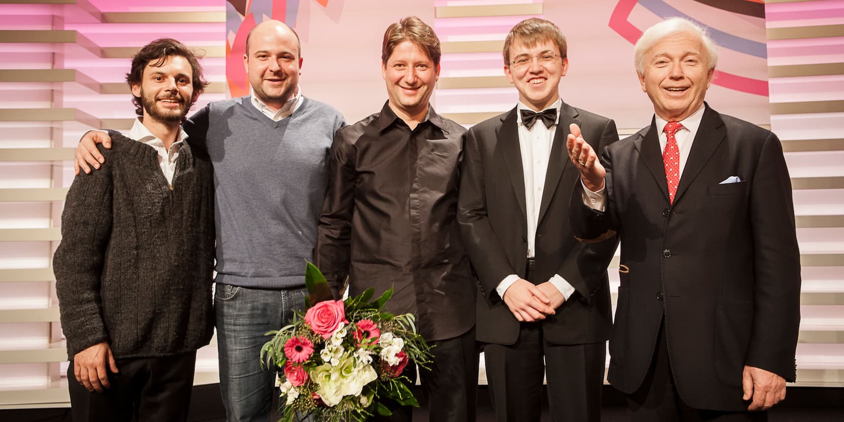 Five men stand side by side, smiling at the camera. The man in the center holds a bouquet of flowers.