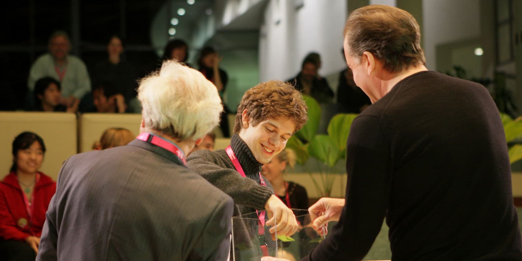 A person is drawing a piece of paper from a glass box, surrounded by two other people. More people are visible in the background.