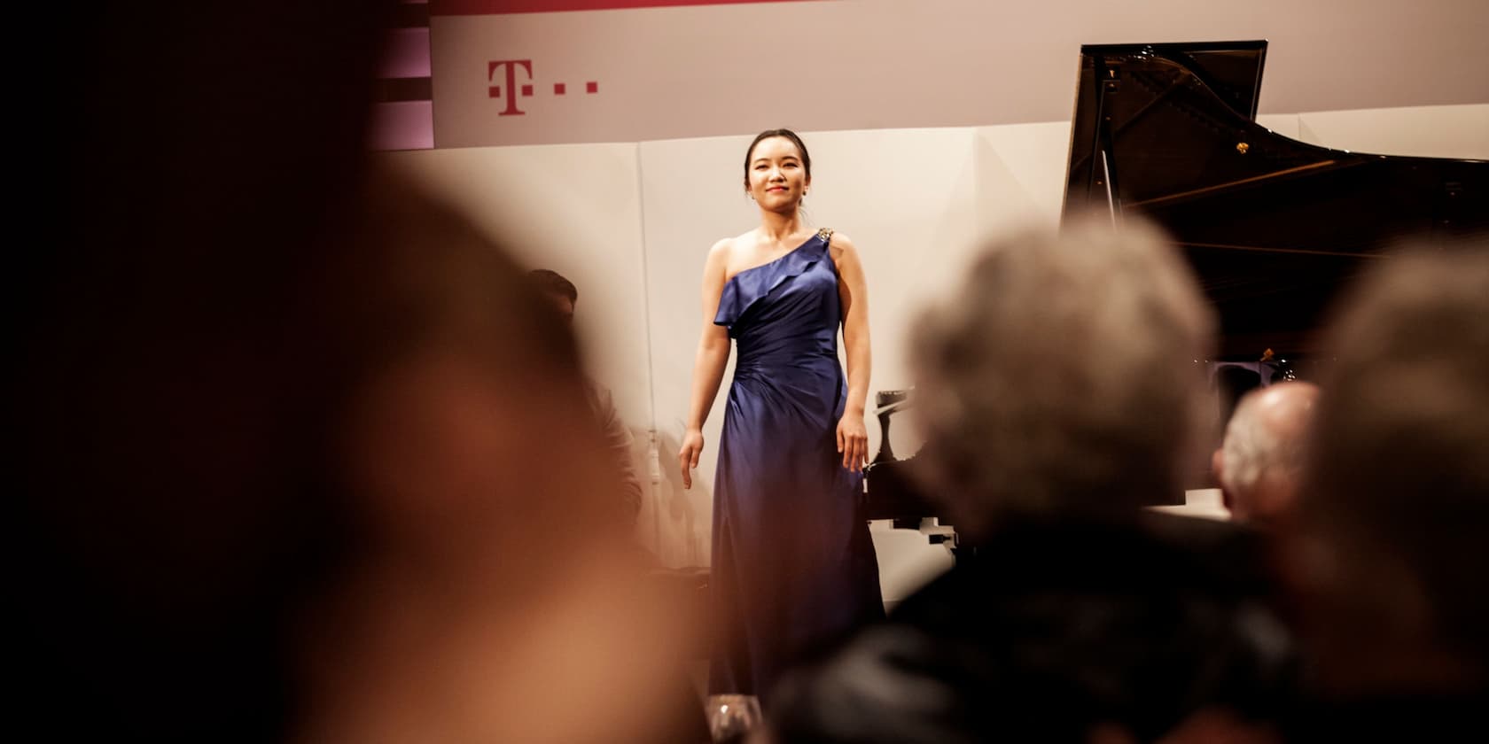 A woman in a blue dress stands on a stage smiling at the audience.