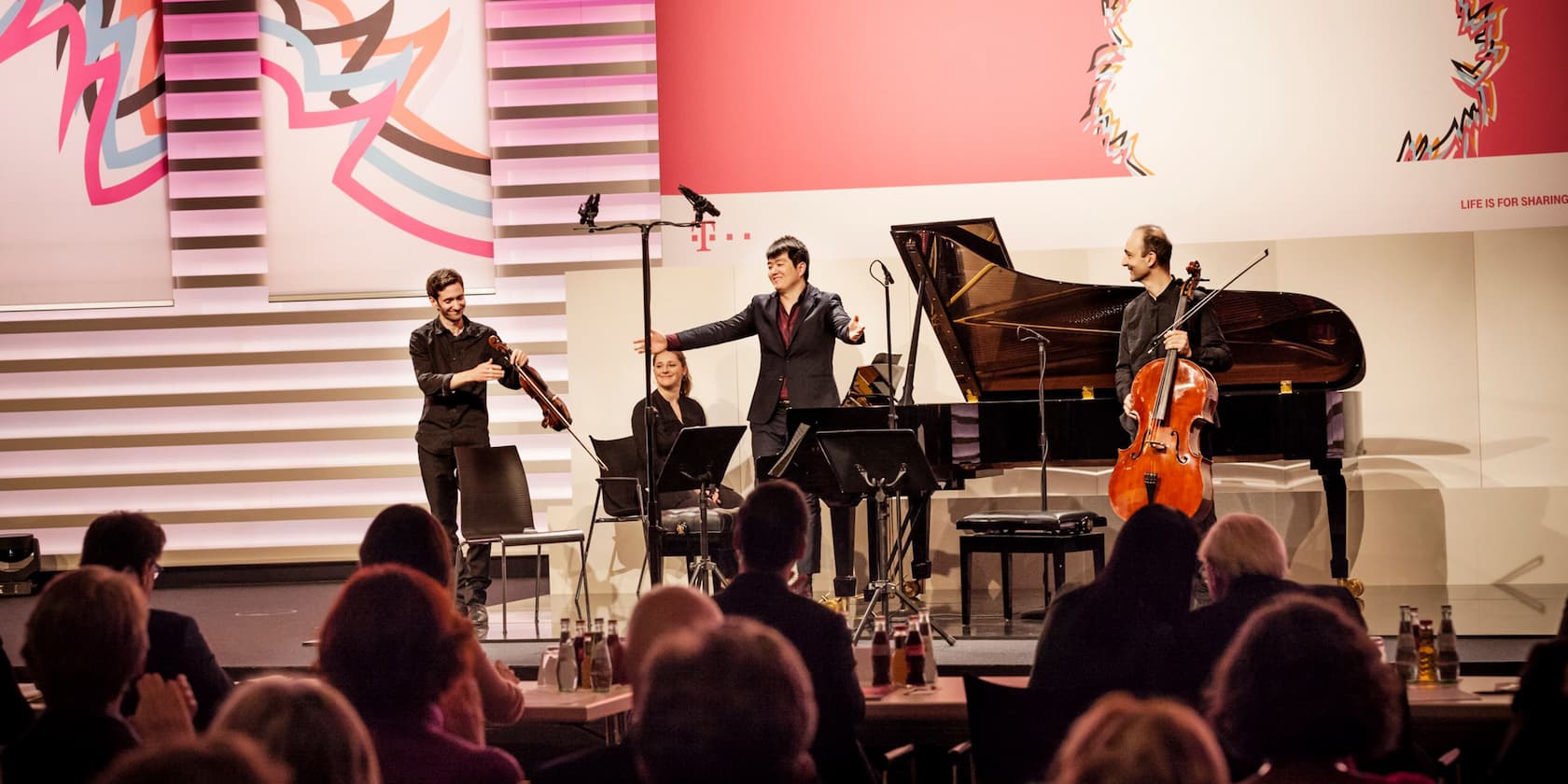 Musicians stand on stage and bow to the audience. A pianist, a cellist, and a violinist are visible.