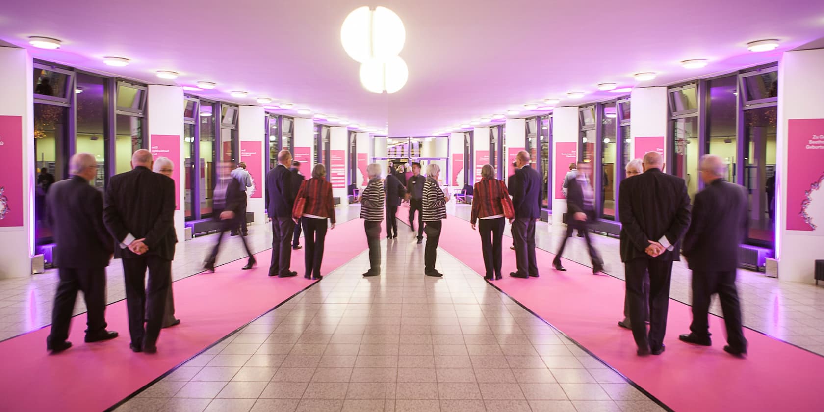 People walking and standing in a corridor with a pink carpet, illuminated by lights.