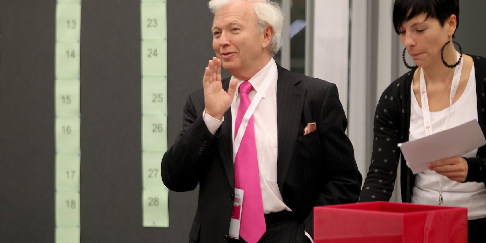 Older man in a suit with a pink tie speaks as a woman stands beside him looking at a document.