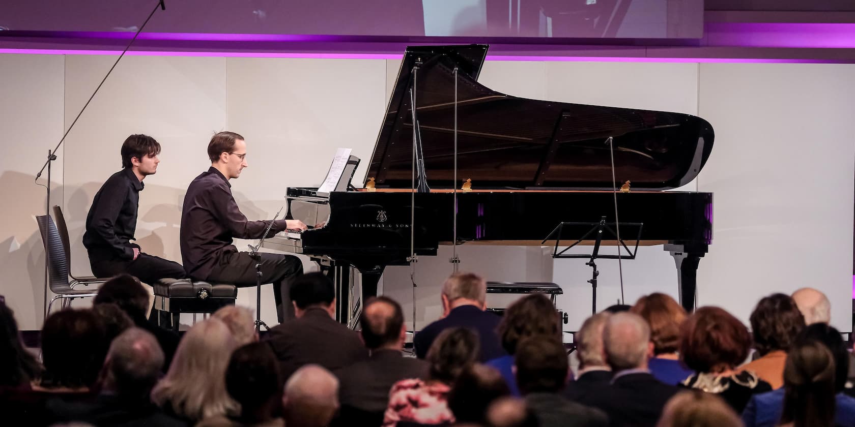 A pianist is playing on a grand piano in front of an audience, with another man sitting next to him.