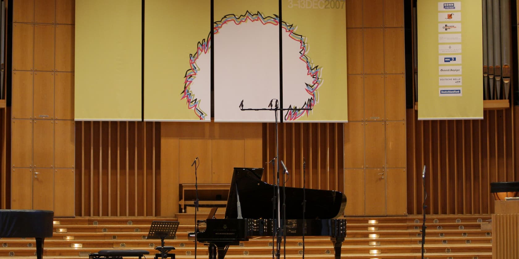 Empty concert hall with a grand piano and several microphones on the stage. A large banner in the background with a creative design and sponsor logos.