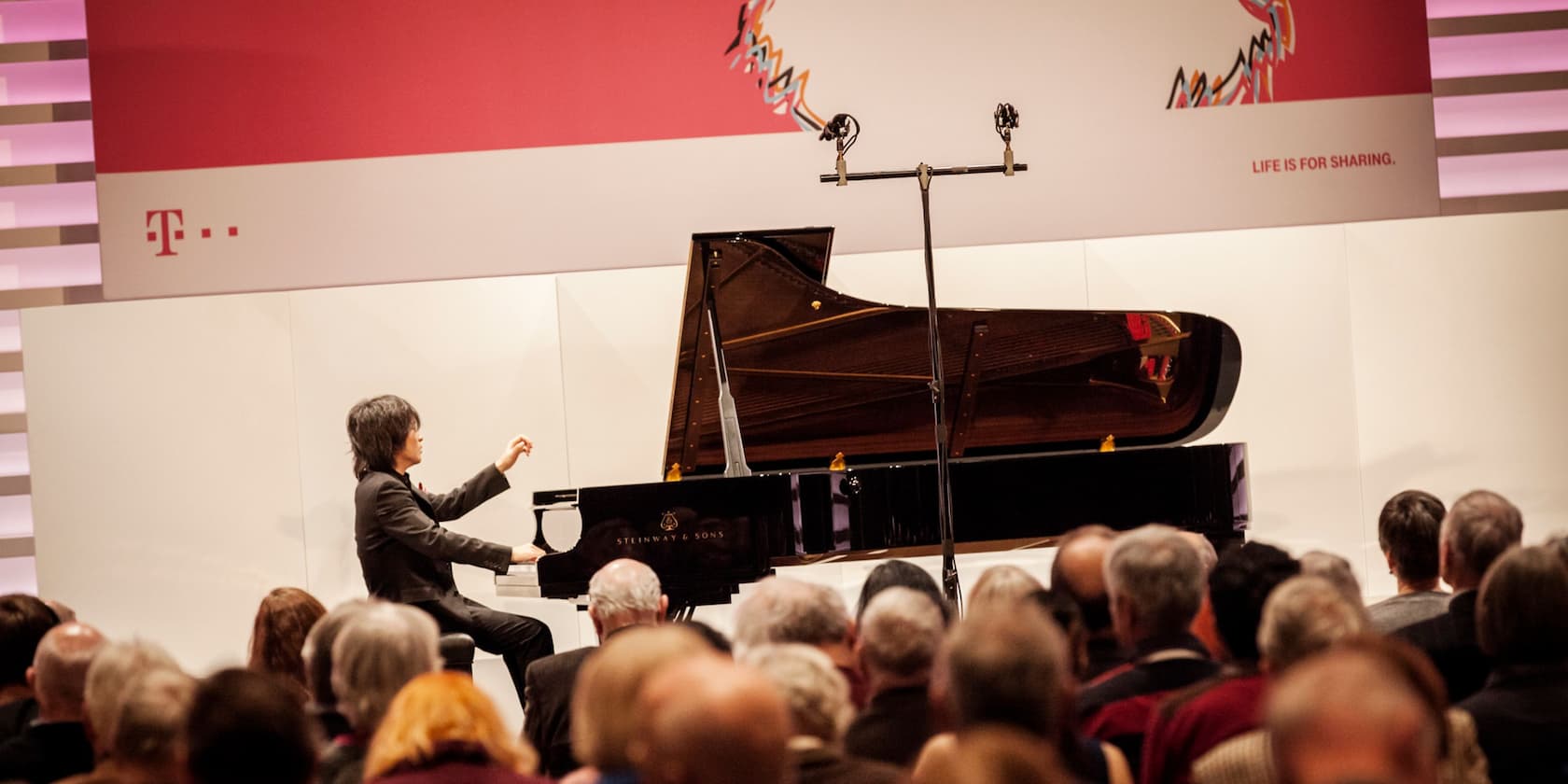 A concert pianist plays the piano on a stage in front of an audience. In the background, a red slogan reads 'Life is for sharing'.