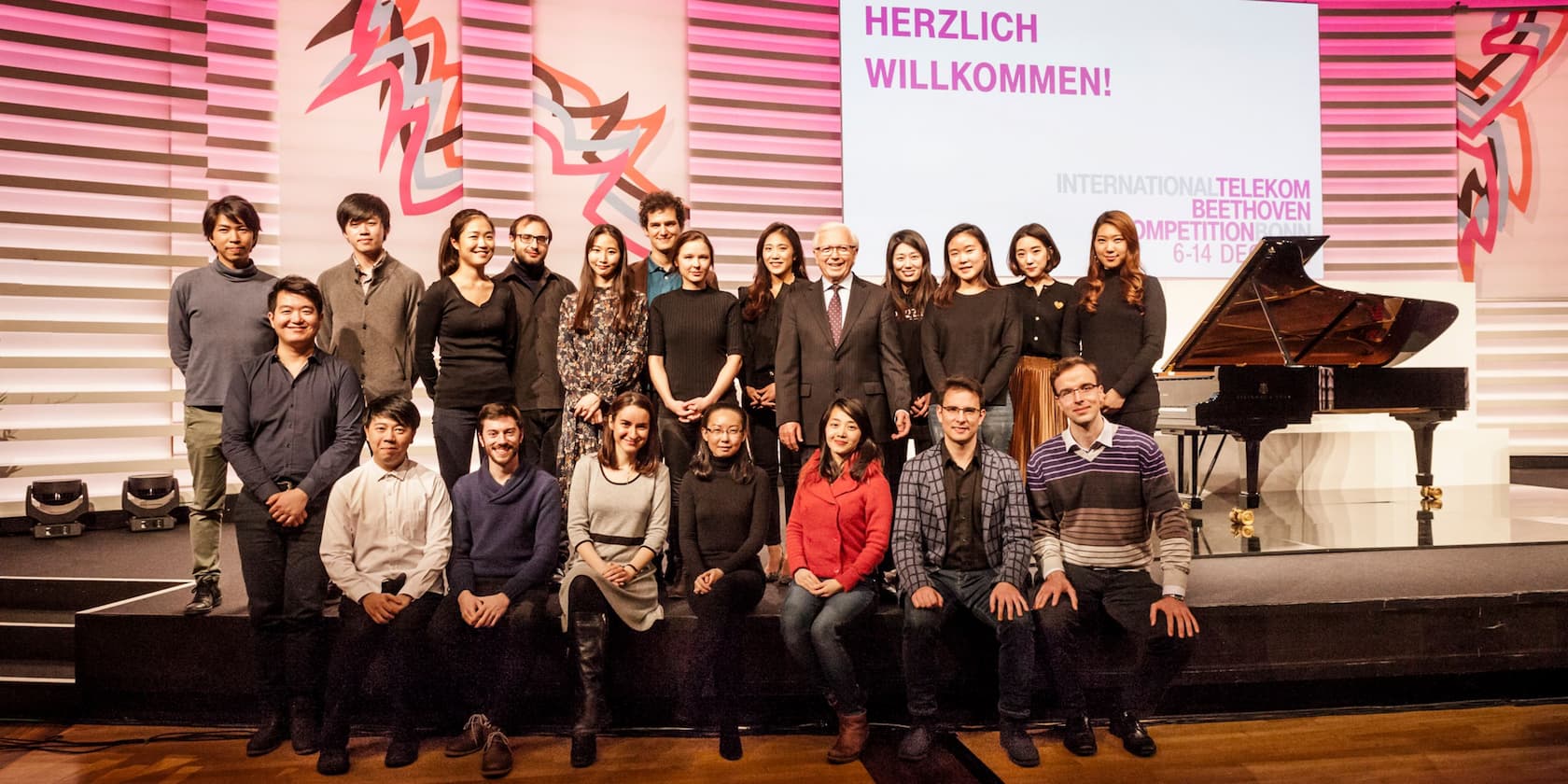 A group of people stand and sit on a stage with a piano in the background. 'Welcome!' is written above.