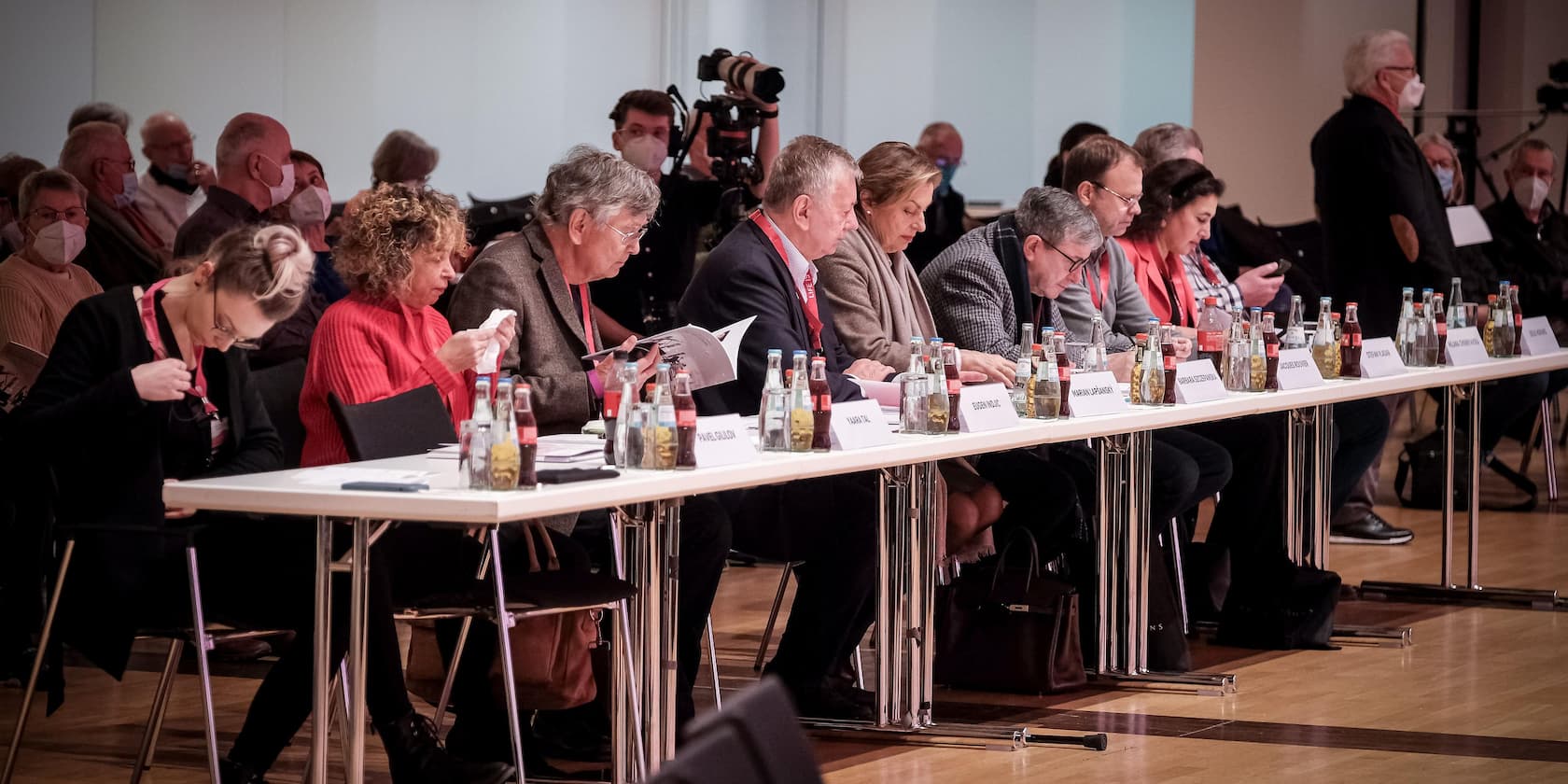 Menschen sitzen in einer Reihe an einem Konferenztisch und lesen Dokumente. Im Hintergrund weitere Teilnehmer und Kameras.