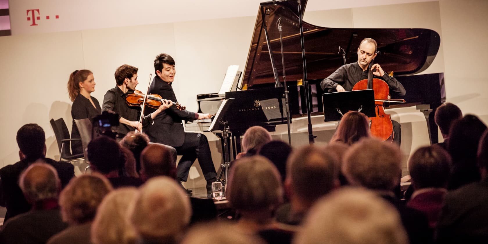 Musicians playing violin, piano, and cello in front of an audience.