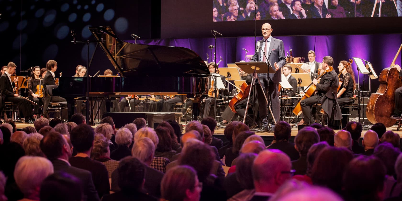 A person speaks on a stage in front of an orchestra during a concert. A grand piano is in the background, with a large audience in the foreground.