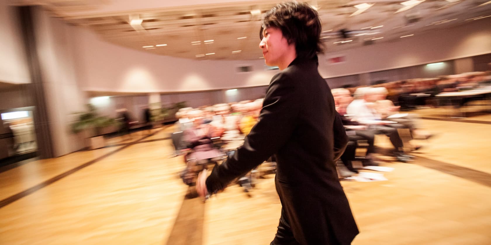 Person in formal attire walking through a conference room with an audience in the background.