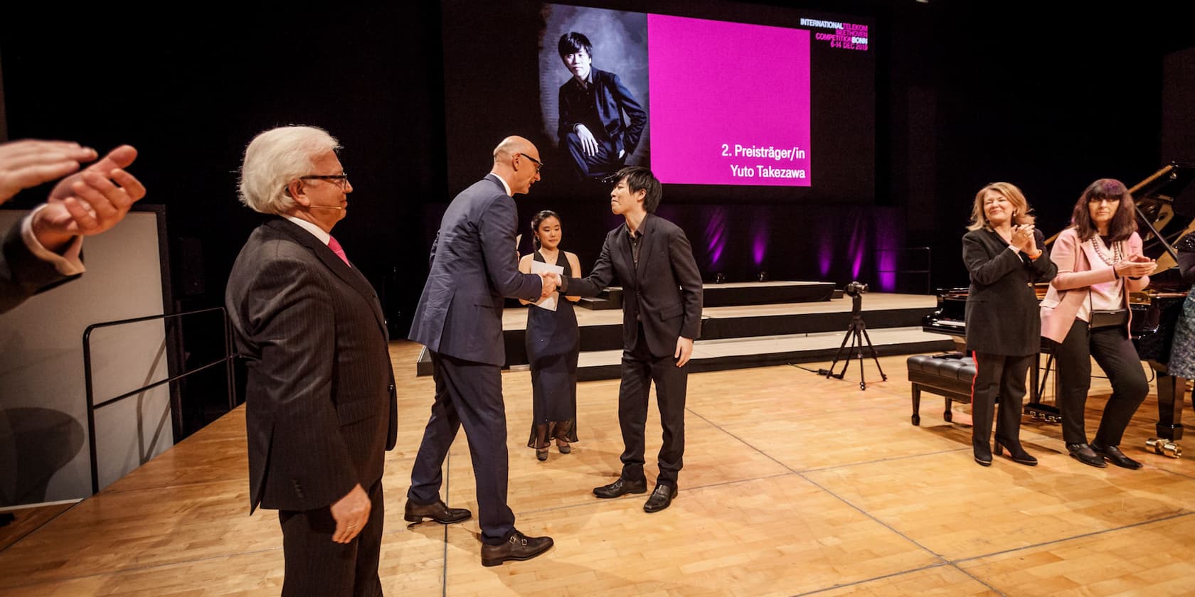 Yuto Takezawa erhält den 2. Preis bei einem Musikpreiswettbewerb und schüttelt die Hand eines Mannes in einem blauen Anzug. Weitere Personen klatschen im Hintergrund.