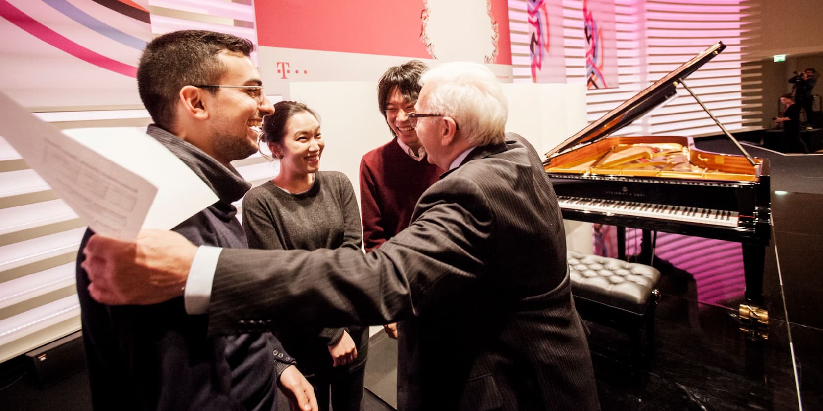 A group of four people laughing and hugging near a grand piano.