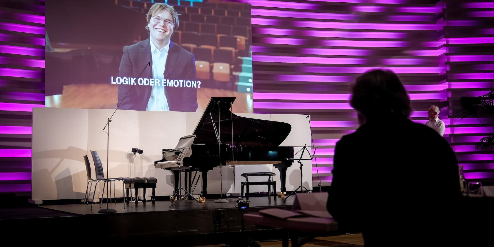 A stage with a grand piano and chairs. In the background, a screen shows a smiling man with the question 'LOGIC OR EMOTION?'.