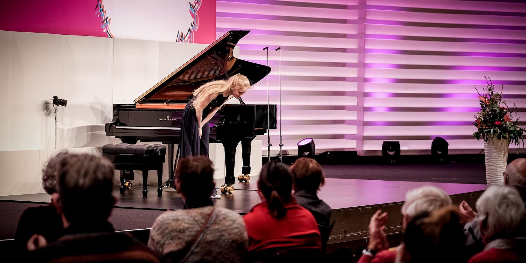 A pianist is bowing in front of a grand piano at a concert while the audience applauds.