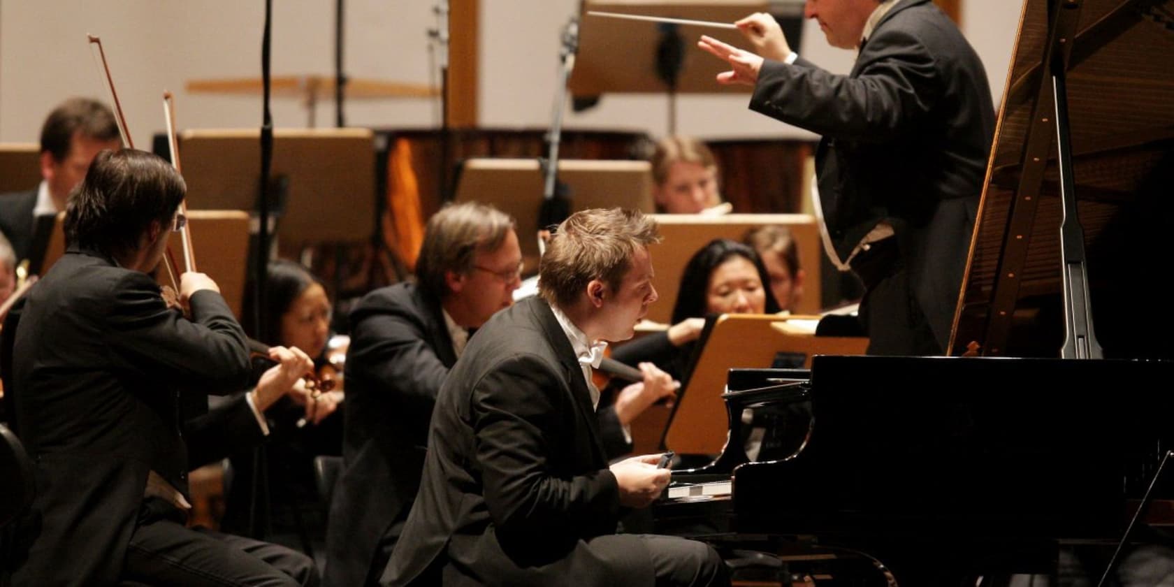 An orchestra performs under the direction of a conductor; a pianist is seated at the piano.