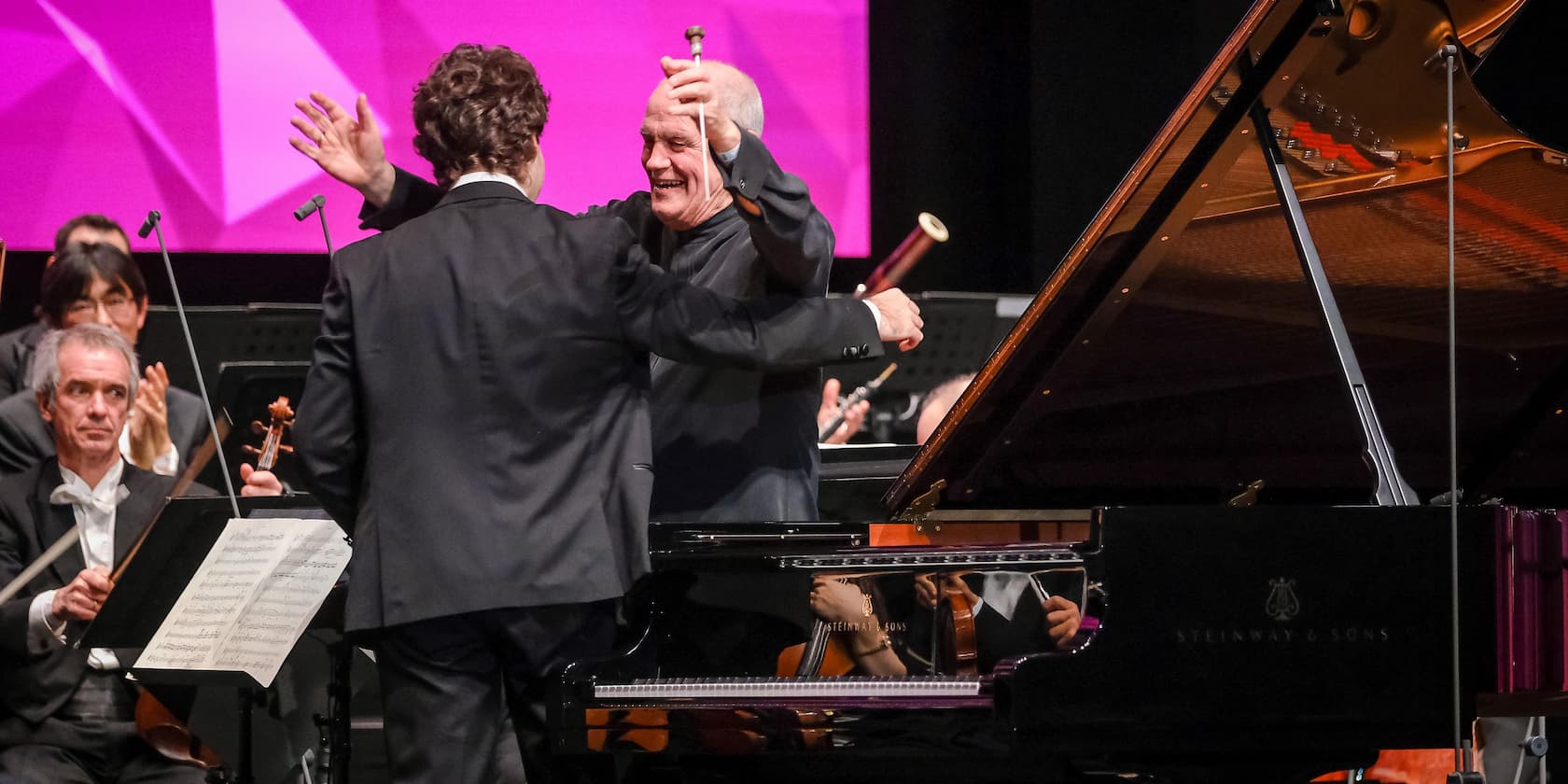 Two musicians hug on a stage, while other orchestra members sit in the background.