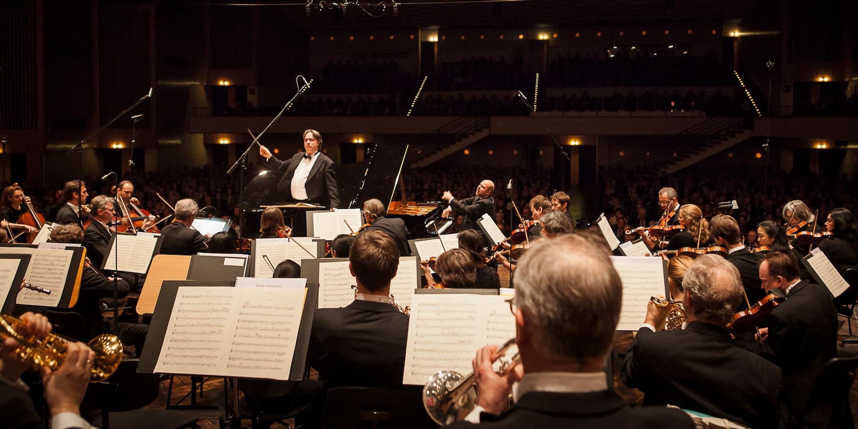 An orchestra performs a concert under the direction of a conductor in a packed concert hall.