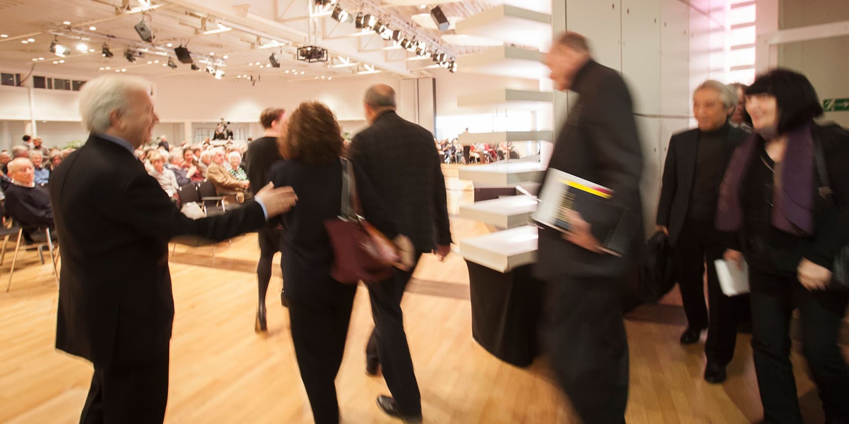 People walk through a room at an event in front of a seated audience.